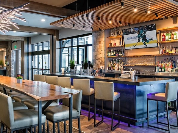 A modern bar area with a wooden bar counter, seating with cushioned chairs, and a large window providing natural light.