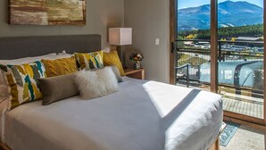 Ein Schlafzimmer mit einem großen Bett, einem Nachttisch mit Lampe und Blick auf eine gebirgige Landschaft durch eine Schiebetür.