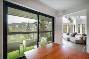 A modern interior with a dining area, a sofa, and a balcony with a view of a forested landscape.