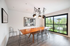 Un comedor moderno con una mesa de madera, sillas traslúcidas, un cuadro abstracto enmarcado y vistas a la vegetación a través de un ventanal grande.