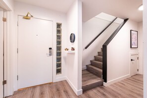 A hallway with a white door, a built-in bookshelf, a clock, and a staircase with wooden steps and a black handrail.