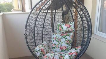 Een hangstoel met bloemenkussens op een balkon met betegelde vloer en een raam.