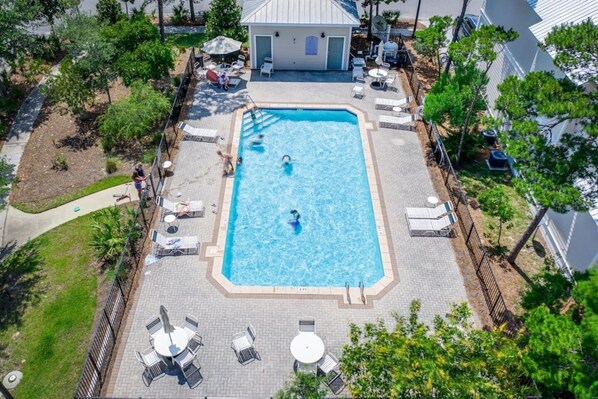 A rectangular swimming pool surrounded by lounge chairs and tables, with a covered seating area and a building in the background.