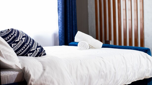 En snyggt bäddad säng med ett vitt täcke och en hoprullad handduk, en mönstrad kudde och en blå sänggavel i ett hotellrum.