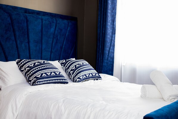 A neatly made bed with white linens and blue patterned pillows, a blue headboard, and a rolled white towel on the side.