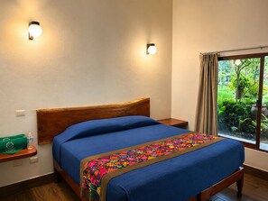 A bedroom with a wooden headboard, a bed with a blue bedspread, a colorful embroidered throw, a nightstand with a green bag and bottle, and a window with a view of greenery. - Hotel Boutique Finca de la Niebla Coatepec (Tlalnelhuayocan)