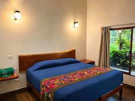 Un dormitorio con cabecera de madera, cama con cubrecama azul, manta bordada colorida, mesita de noche con una bolsa verde y una botella, y una ventana con vistas a la vegetación.