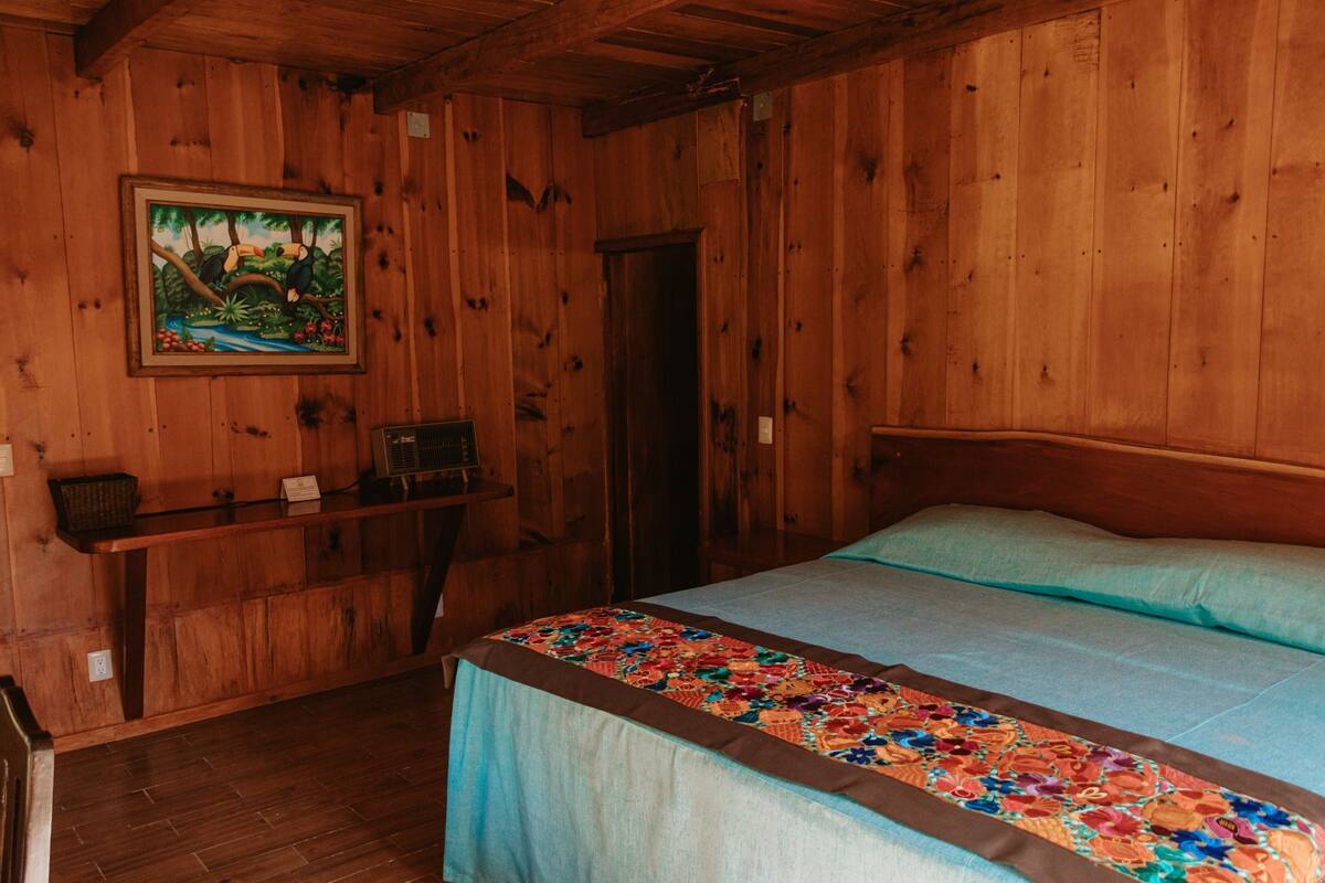 Quarto de madeira com uma cama, uma escrivaninha e um quadro na parede.