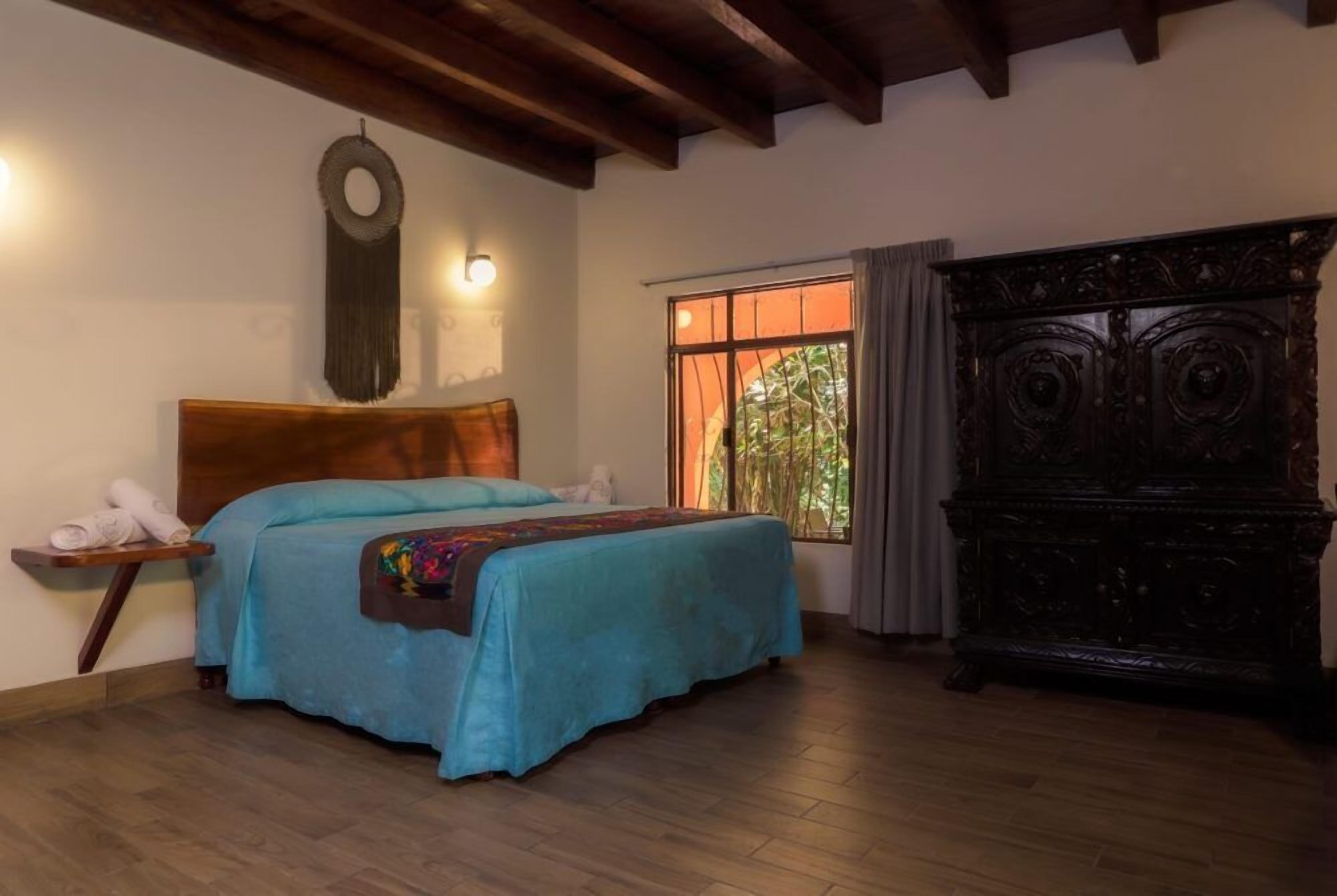 Un dormitorio con cama de madera, una mesita de noche, una lámpara de pared, una ventana con cortinas y un aparador de madera oscura.
