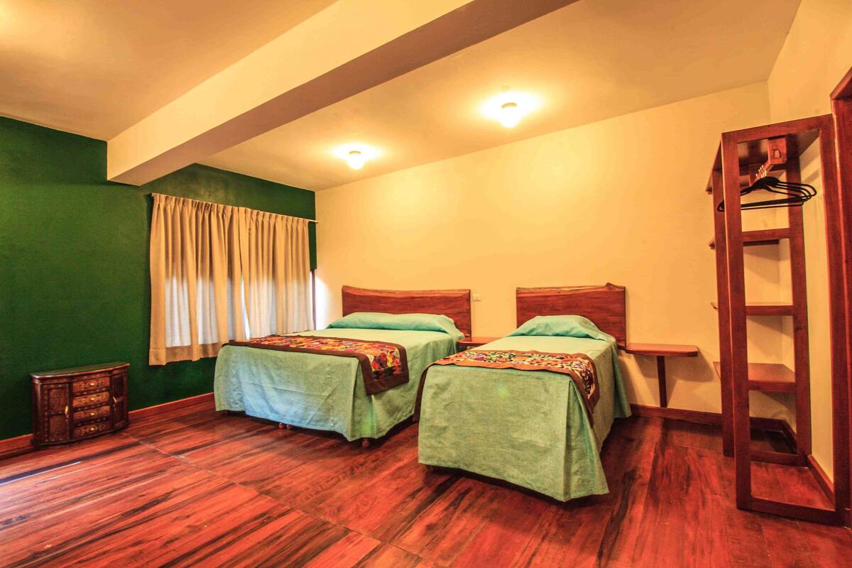 Quarto com duas camas, piso de madeira, janela com cortinas e uma pequena mesa.