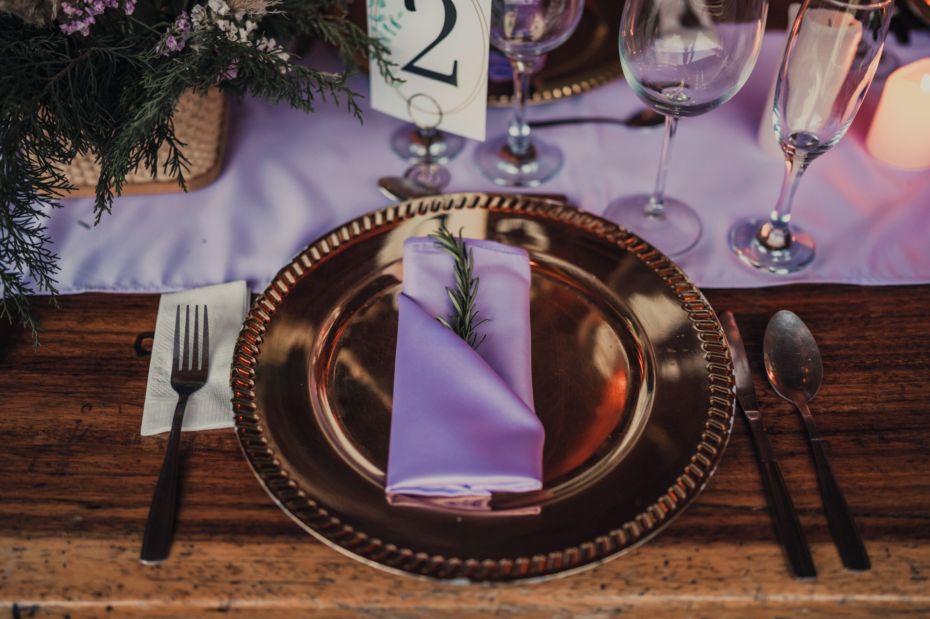 Una mesa dispuesta con un posavasos número 2, una servilleta doblada, una copa, cubiertos y una vela.