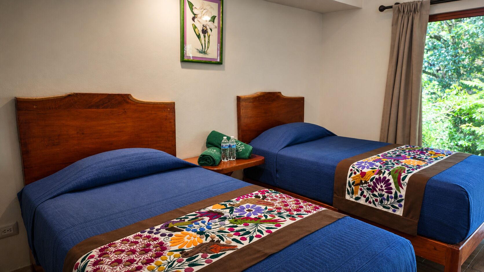 Een hotelkamer met twee bedden, elk voorzien van een blauwe spreidek en een kleurrijke geborduurde deken. Er staat een klein houten nachtkastje met groene handdoeken en waterflessen. Daarboven hangt een ingelijst kunstwerk aan de muur.