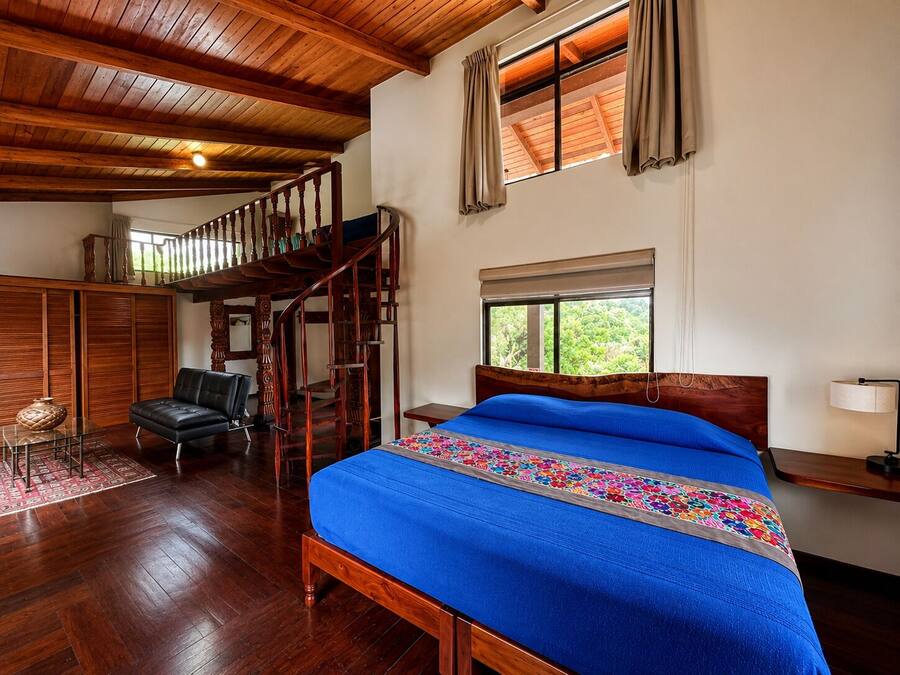 A bedroom with a wooden bed, a staircase, a chair, and a window with curtains.