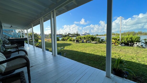 Uma área externa coberta com um deck de madeira, móveis de exterior e vista para uma área gramada com um corpo d'água e edifícios ao longe.