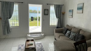 Sala de estar com um sofá bege, tapete estampado e uma pequena mesa de madeira. Há duas janelas com cortinas, vista para um corpo de água e uma porta que dá para o exterior.