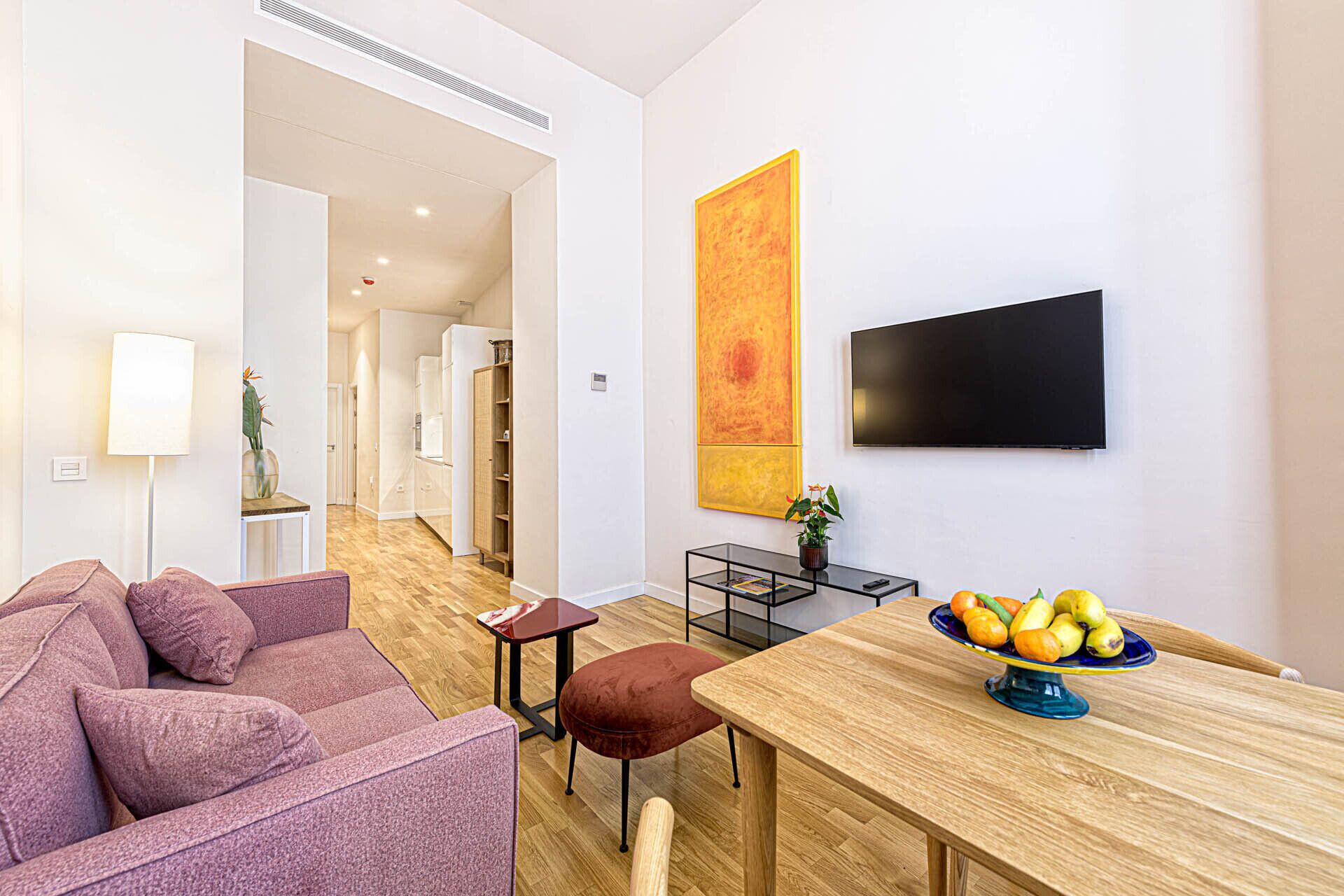 A modern living room with a sofa, a dining table with a fruit bowl, a TV, and a large abstract painting.