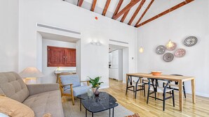 Amplia sala de estar con sofá, una mesa de centro y una mesa de comedor con sillas. Se observa una cocina al fondo, y platos decorativos en la pared.
