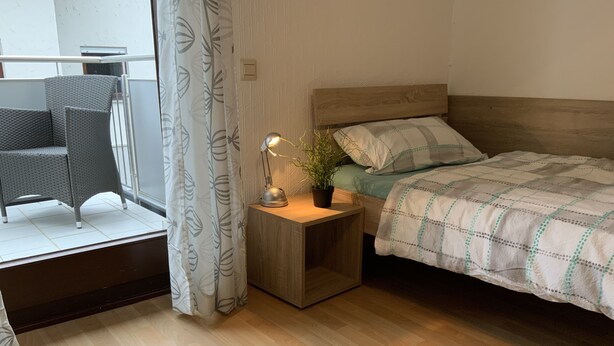 Une chambre avec un lit en bois, une table de chevet avec une lampe, une plante en pot et une chaise près d’une fenêtre ornée d’un rideau.