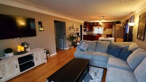 A living room with a large sectional sofa, a TV, and a kitchen in the background. - Charming rental within walking distance to public boat launch on Sage Lake! (Hale)