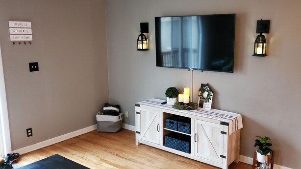 A living room with a flat-screen TV mounted on the wall, two wall-mounted lanterns, a white cabinet with drawers, a small plant, and a sign that reads "There is no place like home." - Charming rental within walking distance to public boat launch on Sage Lake! (Hale)