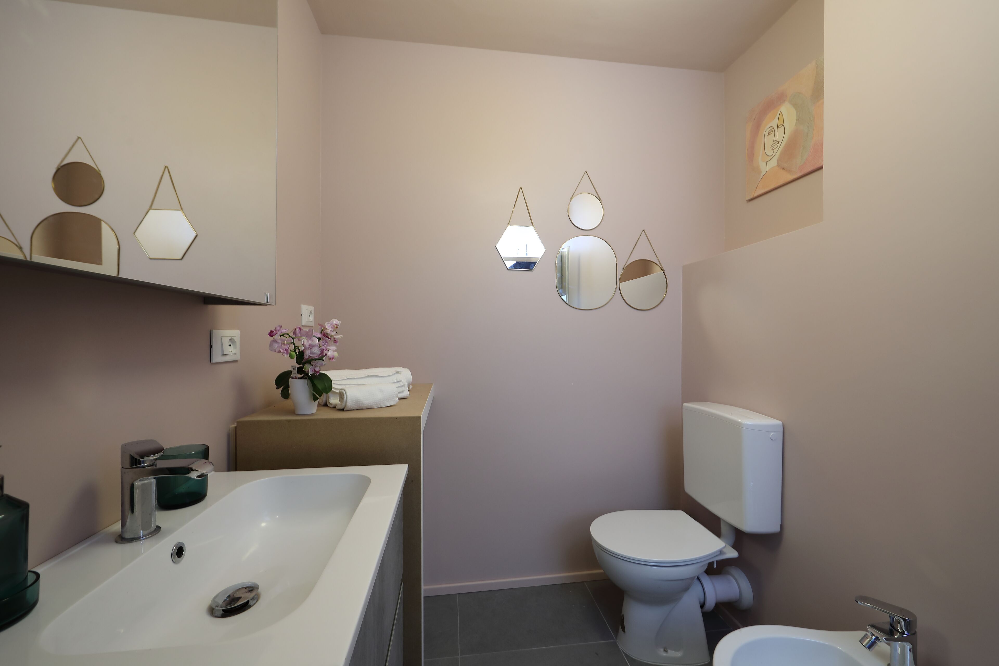 Une salle de bain avec une baignoire, des toilettes et un bidet. On y trouve des miroirs fixés au mur et un vase avec des fleurs sur le comptoir.