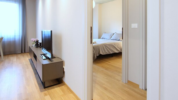 A modern hotel room with a wooden floor, a flat-screen TV on a console, a bed with a white headboard, and a window with curtains. - Brand new apartment in the heart of Udine (Udine)