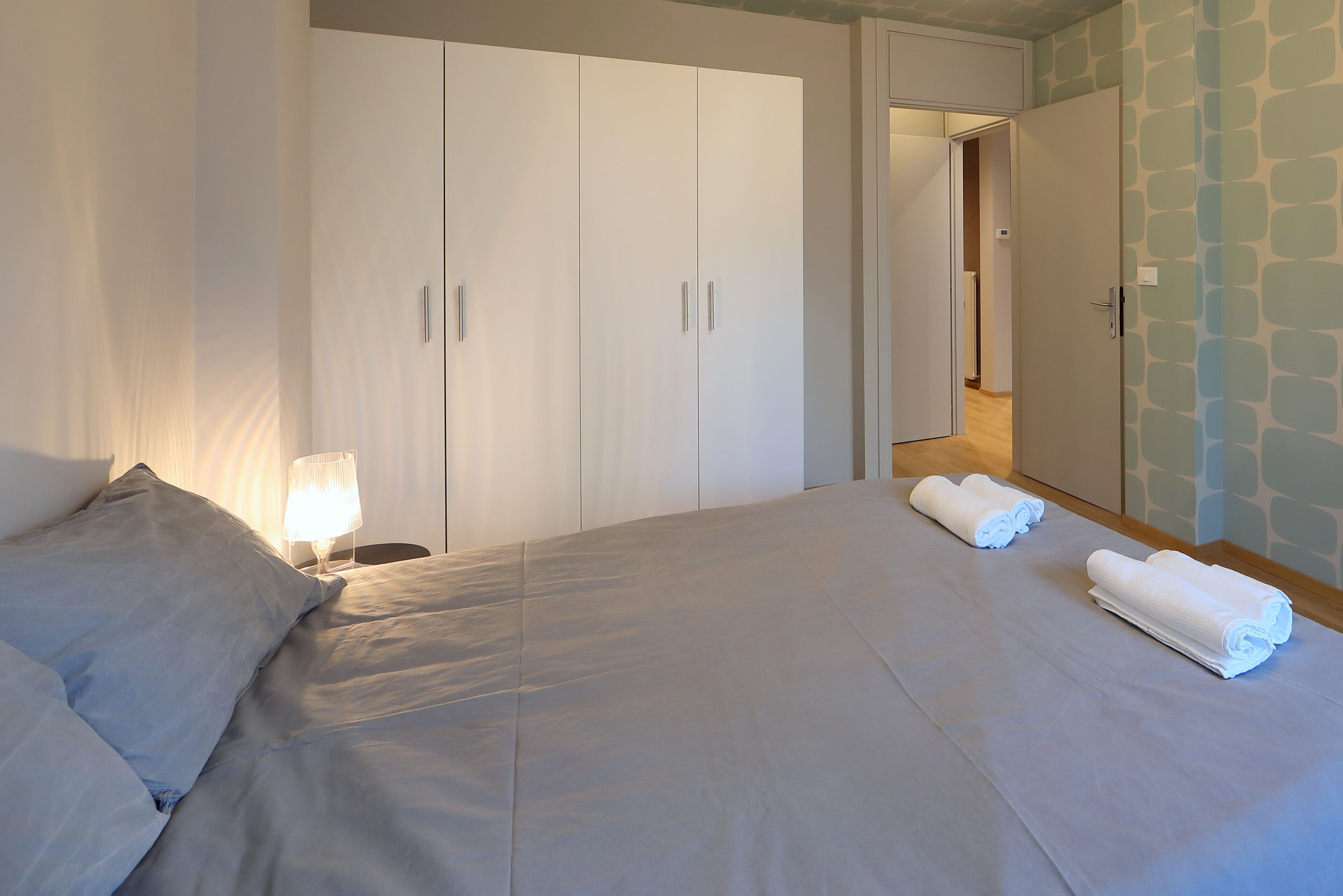 A neatly made bed with white linens and two rolled towels on the bed. There is a nightstand with a lamp beside the bed.