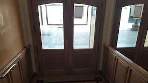 A wooden double door with a black geometric floor design, leading to an outdoor area with a tiled pavement.