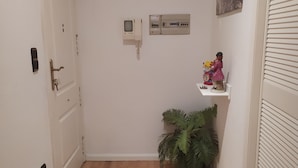 A narrow hallway with a wooden floor, a white door, a wall-mounted intercom, a small shelf with a figurine, and a potted plant.