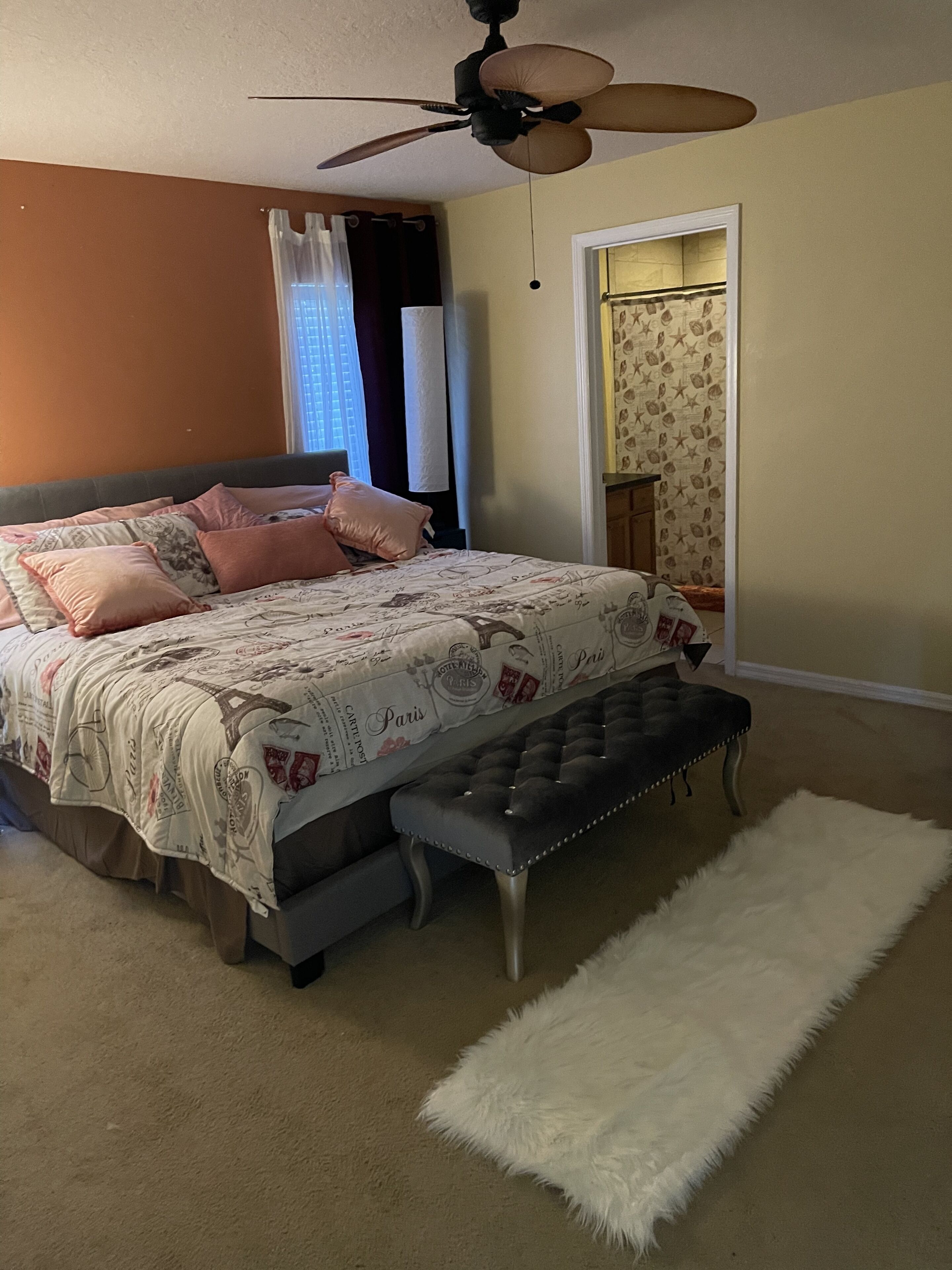 A bedroom with a bed, a tufted bench, a white rug, a ceiling fan, and a door leading to another room.