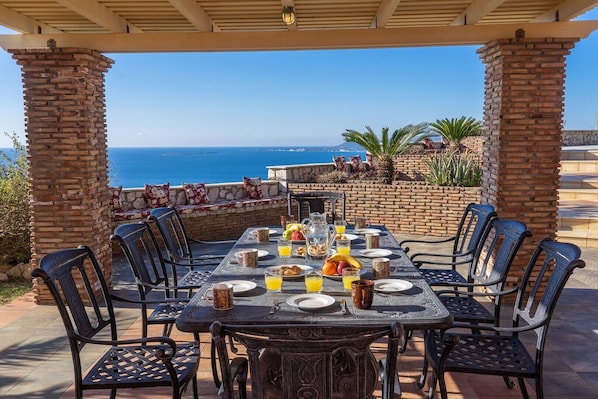 An outdoor dining area with a view of the ocean, featuring a brick column structure, a table set with plates, glasses, and a variety of food and drinks.
