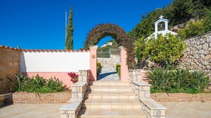 A stone staircase leading to a white building with a bell tower, surrounded by greenery and a clear blue sky. - Sunset Mansion (Argostoli)