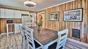 A dining area with a wooden table and chairs, a white ceiling fan, and a kitchen with white cabinets and a microwave. - The Surf Shak-Vintage Coastal Charm (Murrells Inlet)