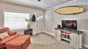 A room with a ceiling fan, a surfboard mounted on the wall, a TV on a wooden stand, a bookshelf, and a patterned sofa. - The Surf Shak-Vintage Coastal Charm (Murrells Inlet)