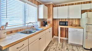 A kitchen with wooden paneling, white cabinets, a stainless steel sink, and a microwave. - The Surf Shak-Vintage Coastal Charm (Murrells Inlet)