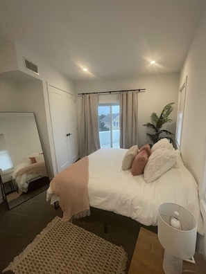 A bedroom with a bed, a bedside table, a lamp, a plant, and a view of the outside.