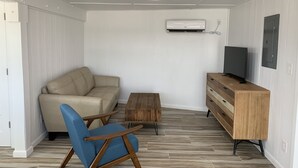 A compact living space with a sofa, a blue armchair, a wooden coffee table, a TV on a wooden cabinet, and an air conditioning unit on the wall.