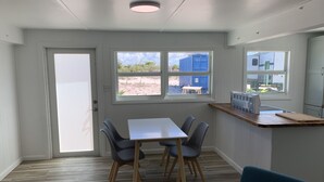 A room with a dining table, four chairs, a bench, a window with a view of the beach, and a kitchenette area.