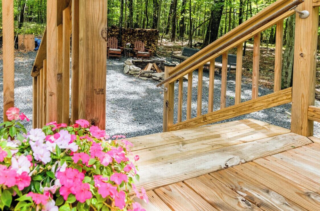 A wooden deck leading to a forested area with a fire pit and chairs.