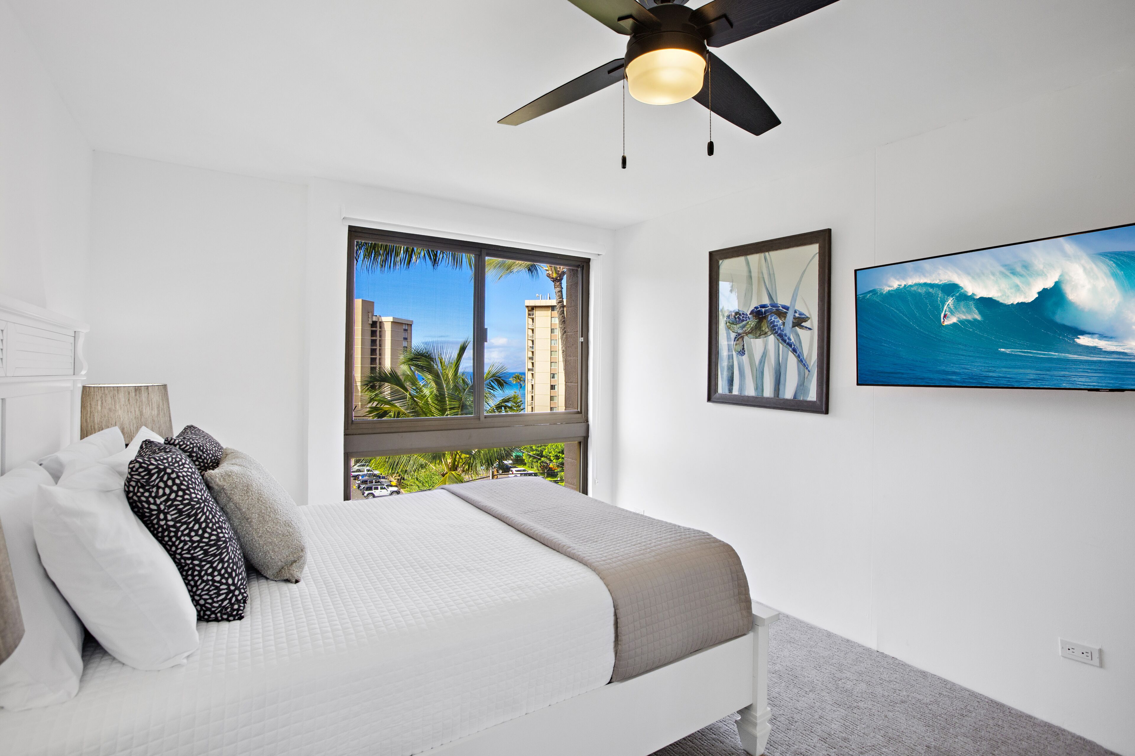 A bedroom with a bed, a ceiling fan, a large window with a view of palm trees and buildings, and a framed picture of a surfer on the wall.
