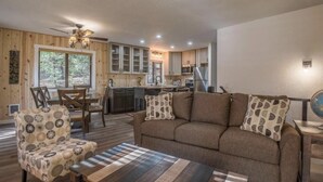 A cozy living room with a sofa, armchair, and coffee table. A kitchen with wooden cabinets and a dining area with a table and chairs are visible. - Modern Cabin w/ EV Car Charger and Pool Access (Dorrington)