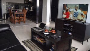 Ein modernes Wohnzimmer mit einem schwarzen Sofa, einem Couchtisch mit einem Laptop, einem Flachbildfernseher und einem Essbereich mit Stühlen und einem Tisch.