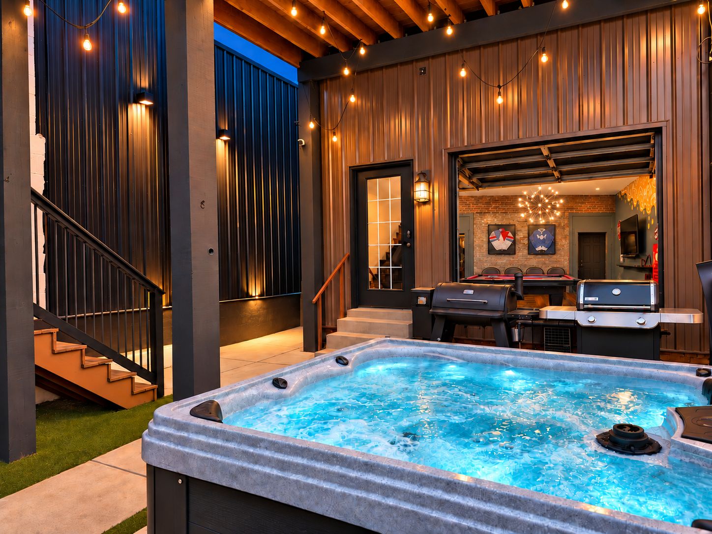 A hot tub in a backyard with a wooden deck, outdoor seating, and a staircase leading to an upper level.