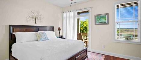 A bedroom with a bed, a ceiling fan, a window with blinds, and a view of greenery outside.