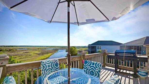 A deck with a glass table and chairs, a grill, and a view of a body of water and landscape. - Your family will be close to everything when you stay at this centrally-located beach oasis (Surf City)