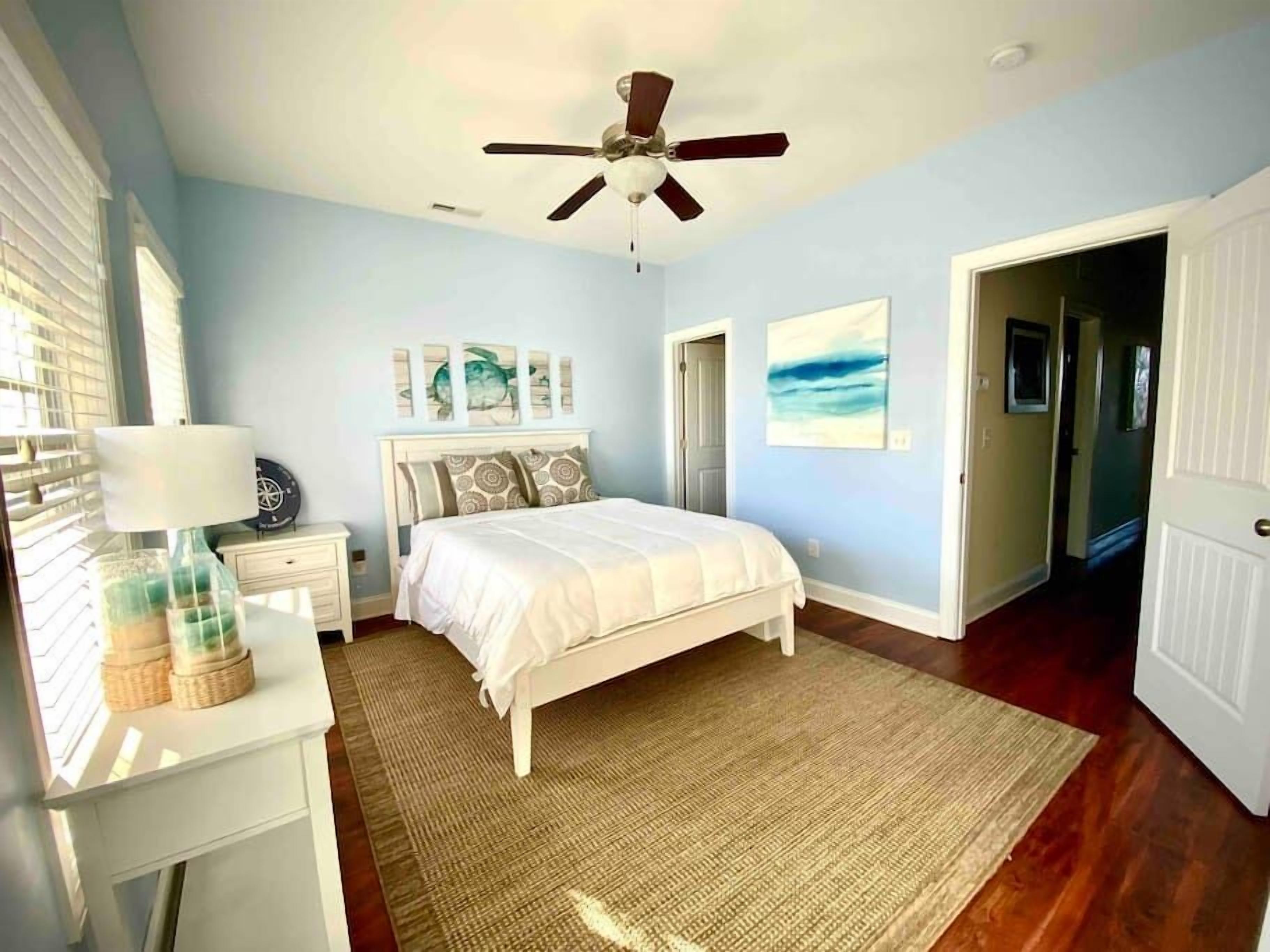 A bedroom with a bed, a nightstand, a lamp, a ceiling fan, and artwork on the walls.