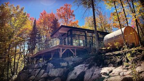 A modern cabin with a glass facade, wooden deck, and a large cylindrical water tank.