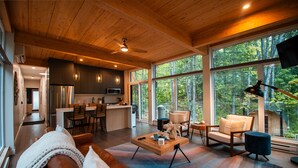 A spacious living area with a wooden ceiling, large windows, a brown leather sofa, a coffee table, and a view of the forest.