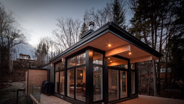 A modern glass house with a wooden deck, surrounded by trees.