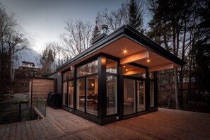 Une maison moderne en verre, dotée d’une terrasse en bois et entourée d’arbres.